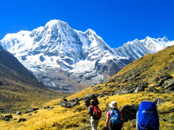 A smiling Nepali woman in traditional clothing stands outside her teahouse, mountains visible in the background.