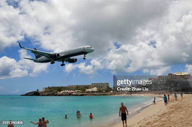 A large commercial airliner flying directly over Maho Beach, with beachgoers silhouetted against the bright sky, conveying the sense of proximity and slightly chaotic energy.