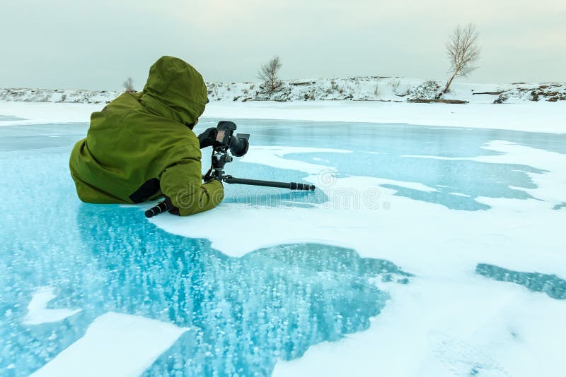 An ice fisherman drills a hole through thick ice on a frozen lake. The image is set during sunrise with golden light and steam coming off the hole, and an angler shown demonstrating the intense cold and serene environment.