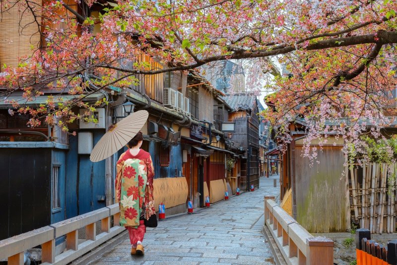 A photo of someone in a kimono standing in front of a traditional wooden building in the Higashiyama district with autumn foliage in the background