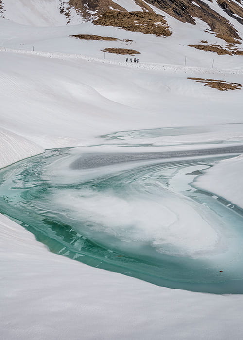 A picturesque winter scene of Bachalpsee Lake, frozen over and covered in snow. The surrounding snow-capped mountains are reflected in the icy surface, creating a stunning mirror effect. A well-maintained hiking trail leads towards the lake.