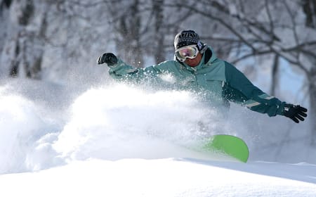 A panoramic shot of a snowboarder riding through pristine powder snow in the Furano backcountry, with the snow-covered peaks of the Daisetsuzan mountain range in the background.