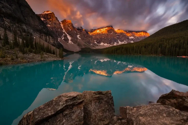 A woman in yoga attire practices yoga outdoors on a wooden platform overlooking the turquoise waters of Lake Louise and the surrounding mountains. The sky is clear blue, and the sun is shining brightly.