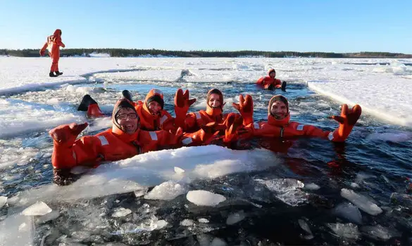 A person in a bright orange insulated suit floating in a dark, icy lake surrounded by snow-covered trees.