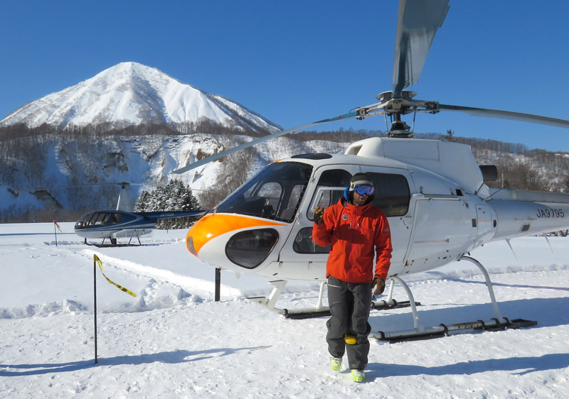 A skier in bright orange gear launching out of a helicopter into deep powder, with Mount Fuji visible in the distance.