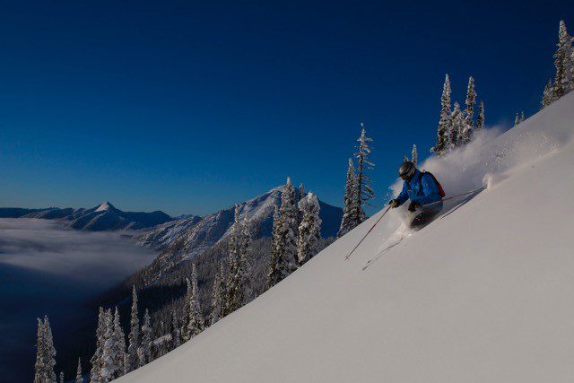 A helicopter lands on a snow-covered mountain with skiers preparing to descend into the untouched powder, showcasing the excitement of heli-skiing in the Monashees.