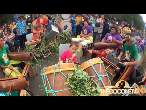 A vibrant scene of drummers performing at Te Maeva Nui in the Cook Islands. The drummers are captured with a motion blur effect, emphasizing their fast movements. Traditional costumes and the ambient dusk light enhance the cultural atmosphere.