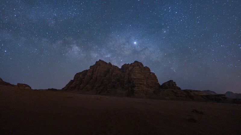 A stunning view of the night sky above the NamibRand Nature Reserve, with the Milky Way arching across the horizon and the silhouettes of desert landscapes in the foreground.