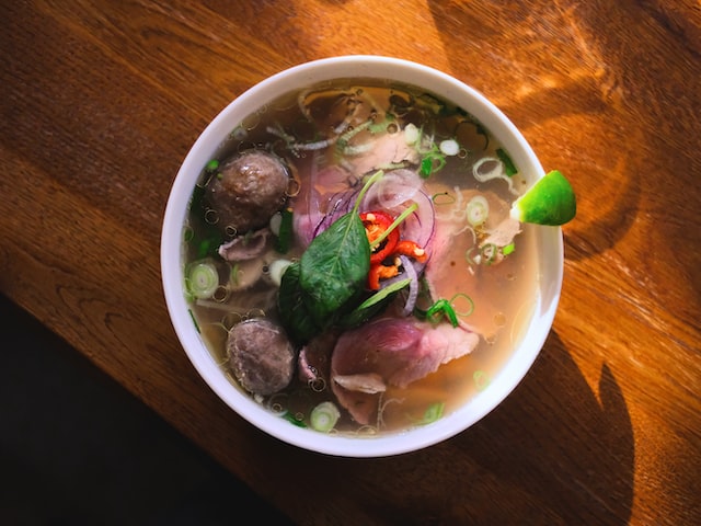 A close-up shot of Bun Bo Hue, showcasing the thick rice noodles, slices of beef, pork knuckle, and vibrant orange chili oil swirling in the broth.