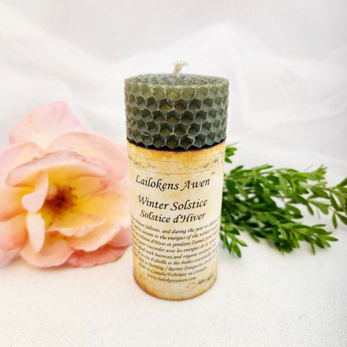 A collection of rustic, hand-poured beeswax candles in various sizes and shapes, made with different foraged herbs, sitting on a wooden shelf.