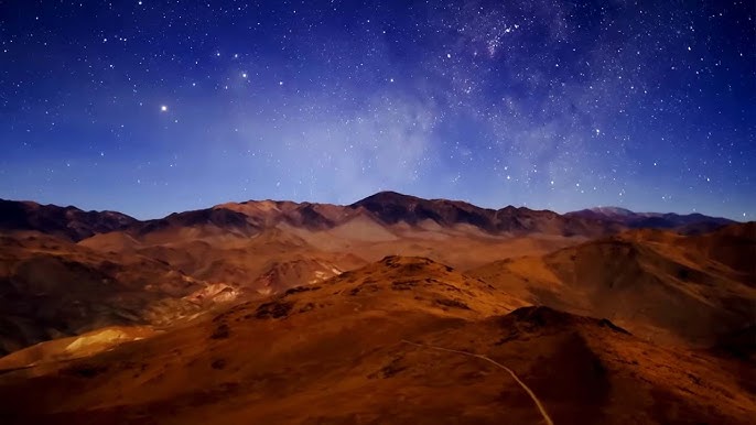 Wide-field astrophotography image of the Milky Way arching over a desert landscape. The sky is filled with stars, and the foreground shows the arid terrain.