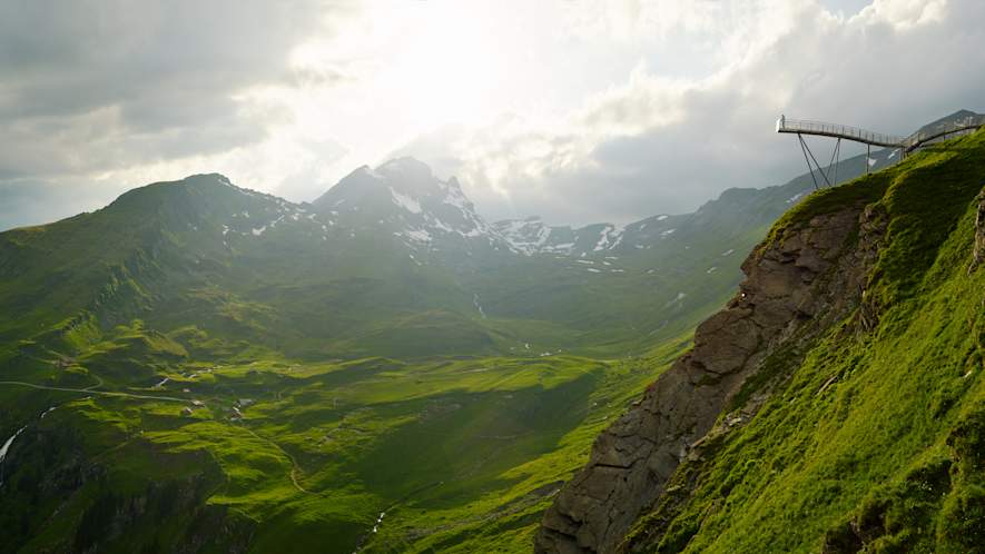 A thrilling view of the Grindelwald First Cliff Walk, a suspended walkway clinging to the edge of a cliff. The walkway offers panoramic views of the surrounding snow-covered landscape and mountains. People are seen walking along the walkway, experiencing the adrenaline rush and the stunning views.