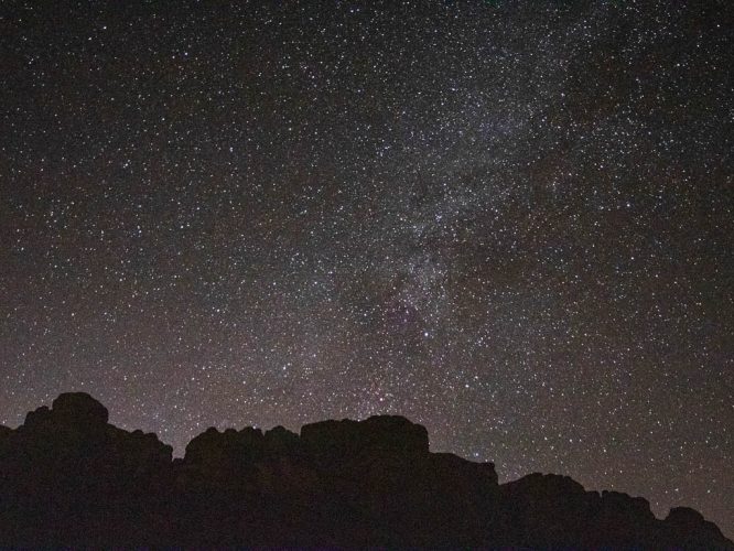 Silhouettes of ancient dunes in the Namib Desert against the backdrop of the Milky Way, highlighting the vastness and tranquility of the Namibian night sky. An Oryx stands majestically in the foreground.