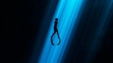 A freediver descends into a sunlit cenote, showcasing the intricate cave system.