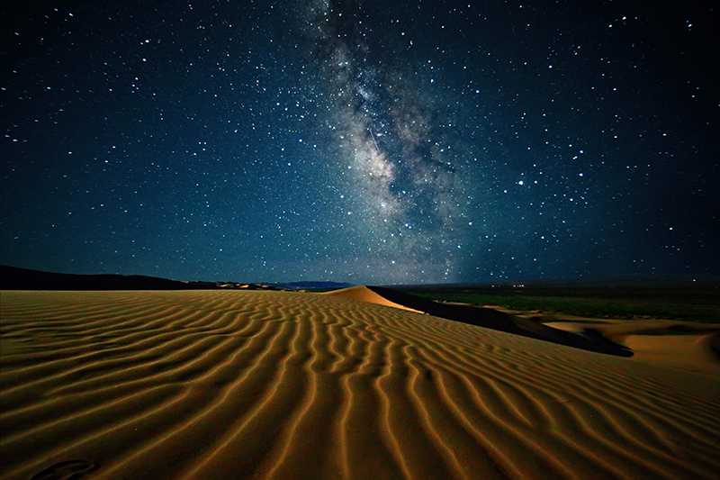 A time-lapse photograph of a traditional ger silhouetted against the swirling Milky Way in the dark Gobi night. The image conveys a sense of solitude, resilience, and the deep connection between the nomadic people and the cosmos.