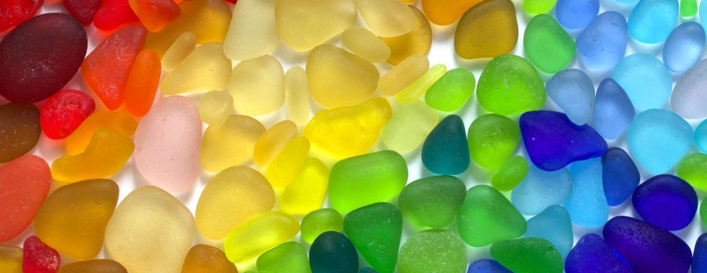 A wide shot of glass beach. Sunlight glints off millions of pieces of smooth, colorful sea glass covering the shoreline. Waves gently lap the shore.