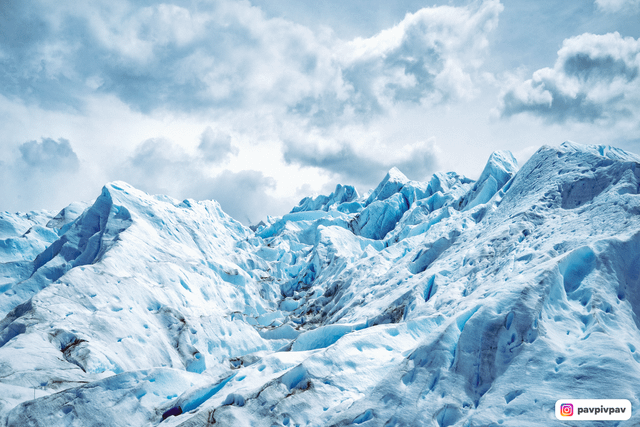 A wide-angle shot of the Perito Moreno Glacier, showing the vastness of the ice field, with a couple silhouetted against the blue ice, wearing crampons and holding ice axes.