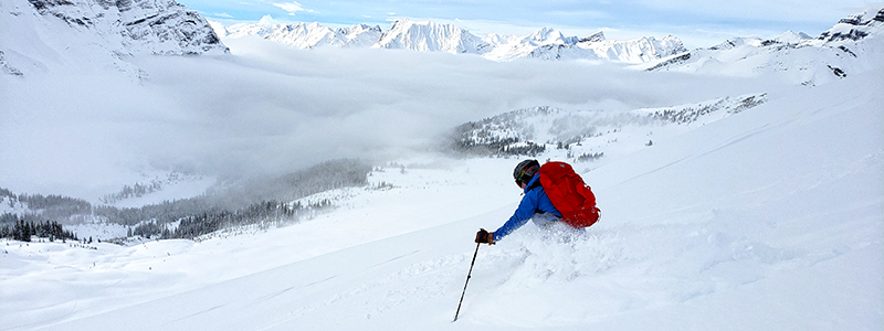 A skier ascending a snow-covered slope with touring gear and a backpack in the Canadian Rockies.