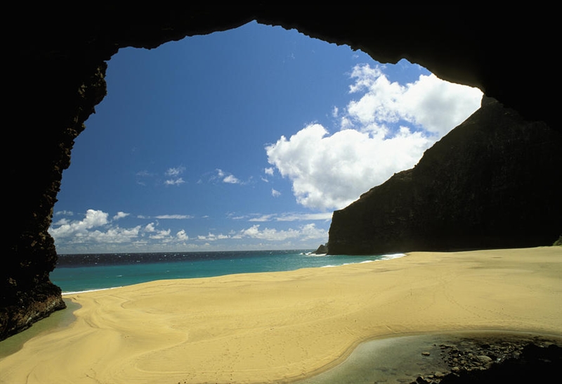 A kayaker paddles through turquoise waters towards the dramatic archway leading to Honopu Beach, bathed in soft light, showcasing the unique access and natural beauty.