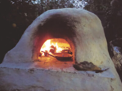A partially completed cob oven dome under construction, showing the layering of the cob and the wooden form used to create the arch. The oven is set against a backdrop of desert scrub and distant mesas. A shovel rests against the base