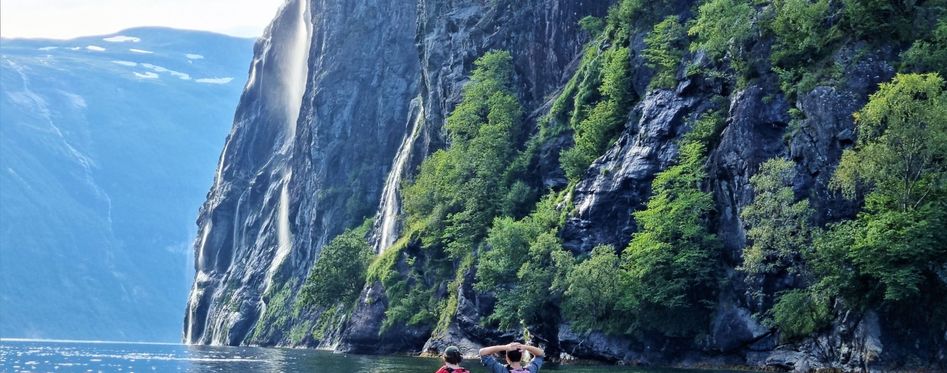Kayakers paddling through the calm waters of a Norwegian fjord, surrounded by towering cliffs and cascading waterfalls.