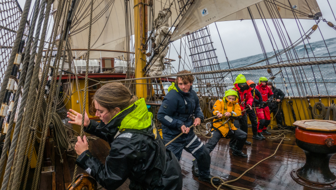 The R. Tucker Thompson under full sail against a backdrop of turquoise waters and lush green islands. The lighting is bright and sunny, with long shadows stretching across the deck.