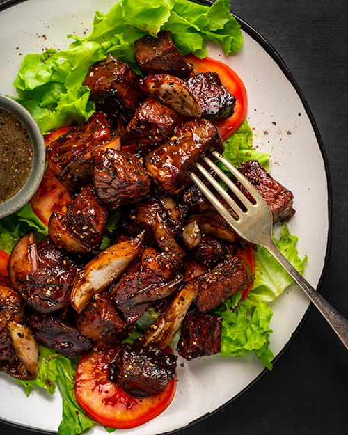 A close-up photo of a Lok Lak dish served on a simple white plate, showcasing the glistening sauce and perfectly cooked beef. The dish is captured under natural light, emphasizing the textures and colors of the ingredients.