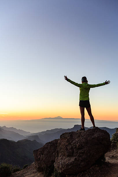 A female mountaineer silhouetted against a vibrant sunrise on a summit, backlit to emphasize her figure against the bright sky. High dynamic range and shallow depth of field enhance the scene.