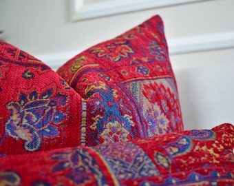 A close-up of three kilim-style pillows in muted earth tones (terracotta, ochre, and dusty rose) arranged on a neutral-colored sofa. The pillows feature geometric patterns inspired by Moroccan and Southwestern designs.