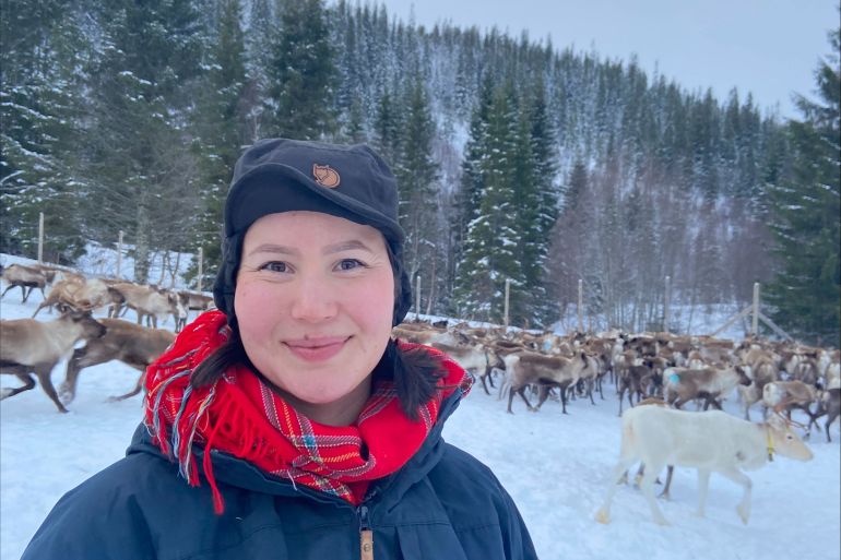 A Sami reindeer herder in traditional clothing stands against a backdrop of a snow-covered landscape, surrounded by his reindeer.