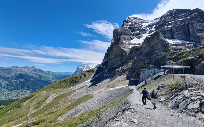 A group of hikers on a trail directly below the Eiger North Face, showing the immense scale of the mountain and the trail's proximity to it.