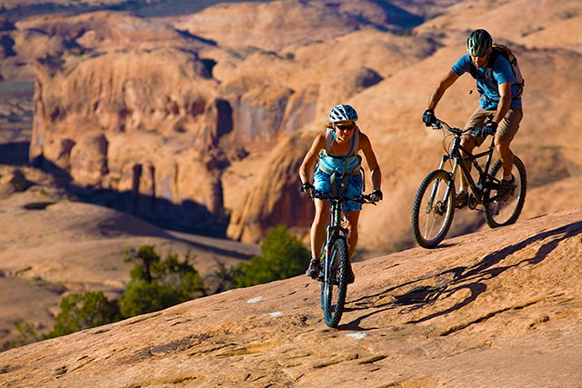 A mountain biker riding the 'Slickrock Trail' in Moab, Utah. The image showcases the iconic red rock landscape, deep blue sky, and the rider's focused determination. The composition highlights the scale of the landscape and the challenging terrain.