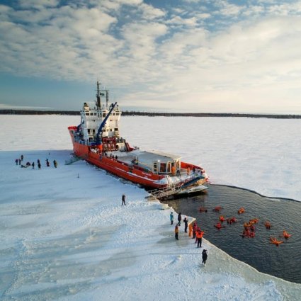 The Icebreaker Sampo cutting through the ice in the Gulf of Bothnia, with people floating in the dark water wearing survival suits, showcasing the adventurous nature of the experience.