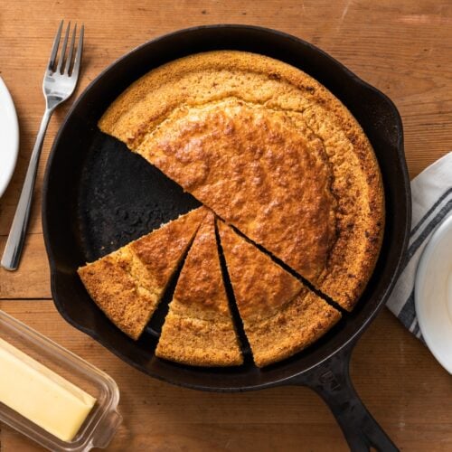 Overhead shot of golden-brown cornbread, baked in a seasoned cast iron skillet, with a dollop of honey butter (slightly melted) on top and a sprig of rosemary. The background includes winter spices such as cinnamon sticks, star anise, and nutmeg arranged artfully on a rustic wooden surface. Warm, inviting lighting mimicking a candle flame, sharp focus on the cornbread.