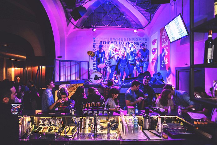 A group of travelers joyfully participating in a pasta-making class at The Yellow hostel, focusing on hands kneading dough with flour dust in the air, and smiling faces all around a large wooden table