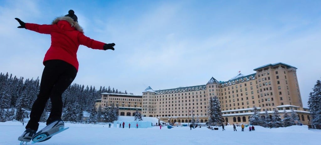 The Fairmont Chateau Lake Louise is majestically reflected in the frozen surface of Lake Louise, surrounded by snow-covered mountains and an ice-sculpture garden. The image emphasizes the winter wonderland atmosphere and the hotel's grand architecture.