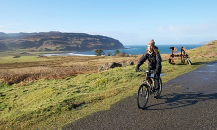 David and Emily cycle past wind turbines on the Isle of Eigg, the rugged beauty of the landscape surrounding them. The clear blue sky accentuates the renewable energy infrastructure. They feel a sense of freedom and adventure as they embrace the vastness of the landscape.