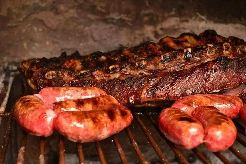 Close-up shot of the asado sizzling on the grill, showcasing the different cuts of meat and their textures. The warm lighting highlights the smoky char and juiciness.