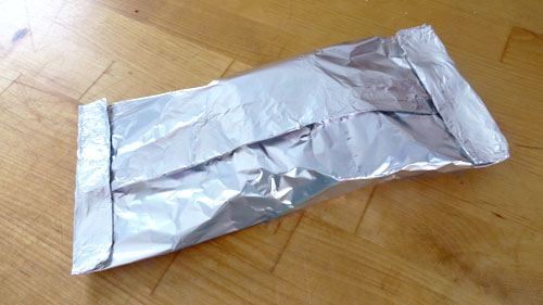 Maya smiling while holding a partially dirty enamel camping mug next to a campfire, pine trees visible in the background