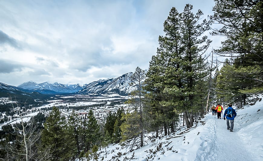 A fat biker pauses on a snow-covered trail with the town of Banff spread out below, showcasing the panoramic views from Tunnel Mountain Trail.